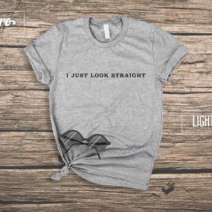 May include: Light grey t-shirt with the text "I just look straight" printed on the front. The t-shirt is laying on a wooden surface with a pair of sunglasses.