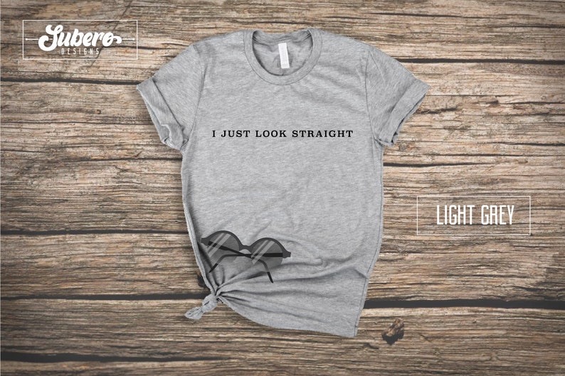 May include: Light grey t-shirt with the text "I just look straight" printed on the front. The t-shirt is laying on a wooden surface with a pair of sunglasses.