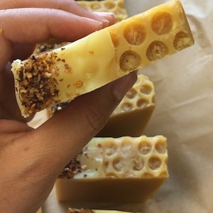 May include: A close-up of a honey-colored soap bar with a honeycomb pattern. The soap bar is being held in a hand and has a layer of brown specks on the bottom.