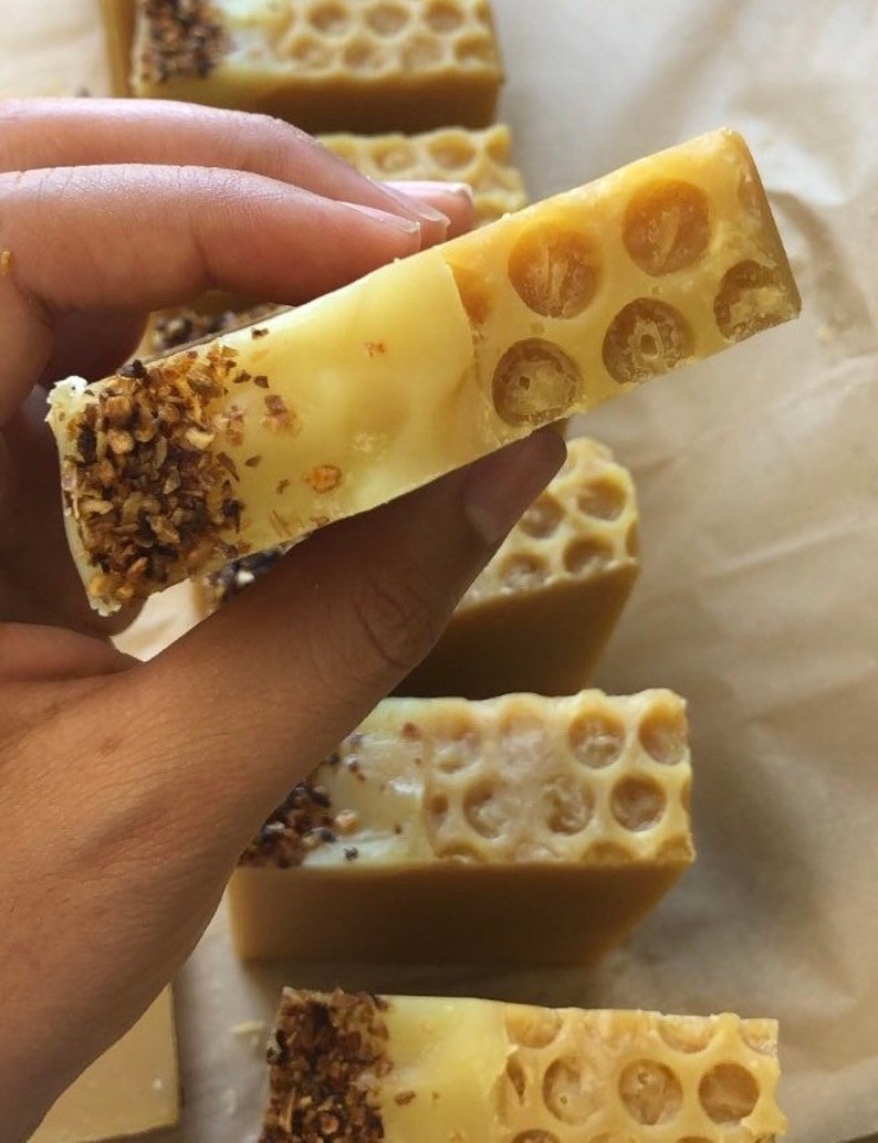 May include: A close-up of a honey-colored soap bar with a honeycomb pattern. The soap bar is being held in a hand and has a layer of brown specks on the bottom.