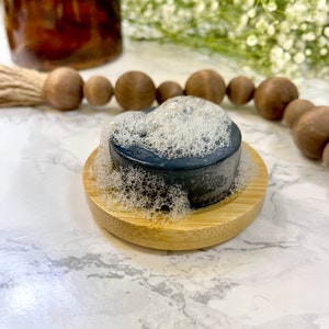 May include: A black charcoal soap bar with white foam on top, sitting on a round wooden soap dish.