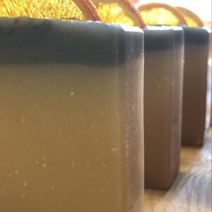 May include: Four bars of handmade soap with a light brown base and a dark gray top. Each bar is topped with a dried orange slice.