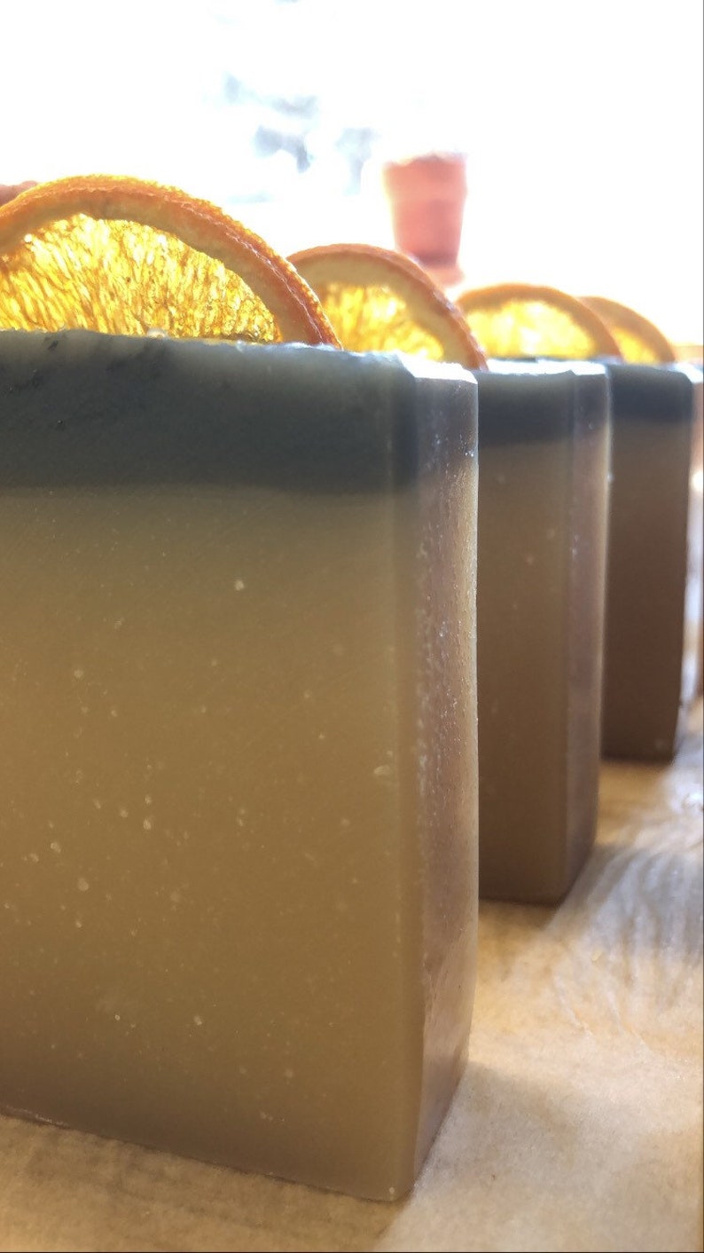 May include: Four bars of handmade soap with a light brown base and a dark gray top. Each bar is topped with a dried orange slice.