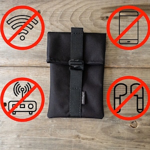 Può includere: Una custodia nera con una tracolla e una chiusura a zip. La custodia è su una superficie di legno con quattro cerchi rossi intorno. I cerchi hanno simboli barrati, tra cui un simbolo wifi, un telefono cellulare, un router e cuffie.