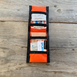 Puede incluir: Botiquín de primeros auxilios naranja con ribete negro, que contiene una variedad de suministros médicos, incluyendo toallitas antisépticas, vendas y una férula. El kit está etiquetado como "SHRUGgear SPLINT"