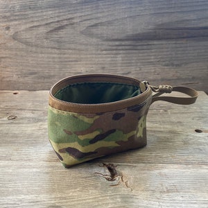 Peut inclure: Une pochette zippée à motif camouflage avec une fermeture éclair marron et une sangle marron. La pochette est ouverte et l'intérieur est doublé de tissu vert.