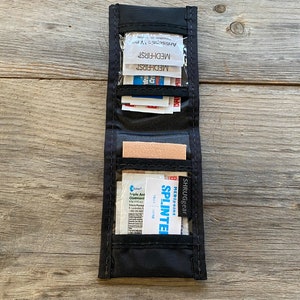May include: Open black nylon first aid kit showing medical supplies. Contents include antiseptic wipes, bandages, and a splinter removal tool. The kit is designed to be compact and portable, ideal for travel or outdoor use.