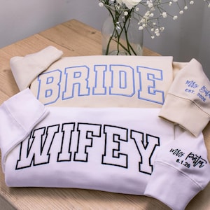 May include: Two sweatshirts, one cream and one white, with embroidered text. The cream sweatshirt says "BRIDE" in blue, and the white one says "WIFEY" in black. Sleeves are embroidered with names and dates. Wedding theme.