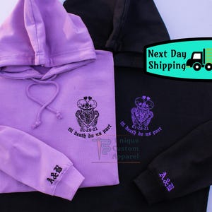 Two hoodies, one black and one lavender, are shown folded with the front of the black hoodie and the back of the lavender hoodie visible. Both have an embroidered design of two skeletons embracing with a heart above their heads, and the text "til