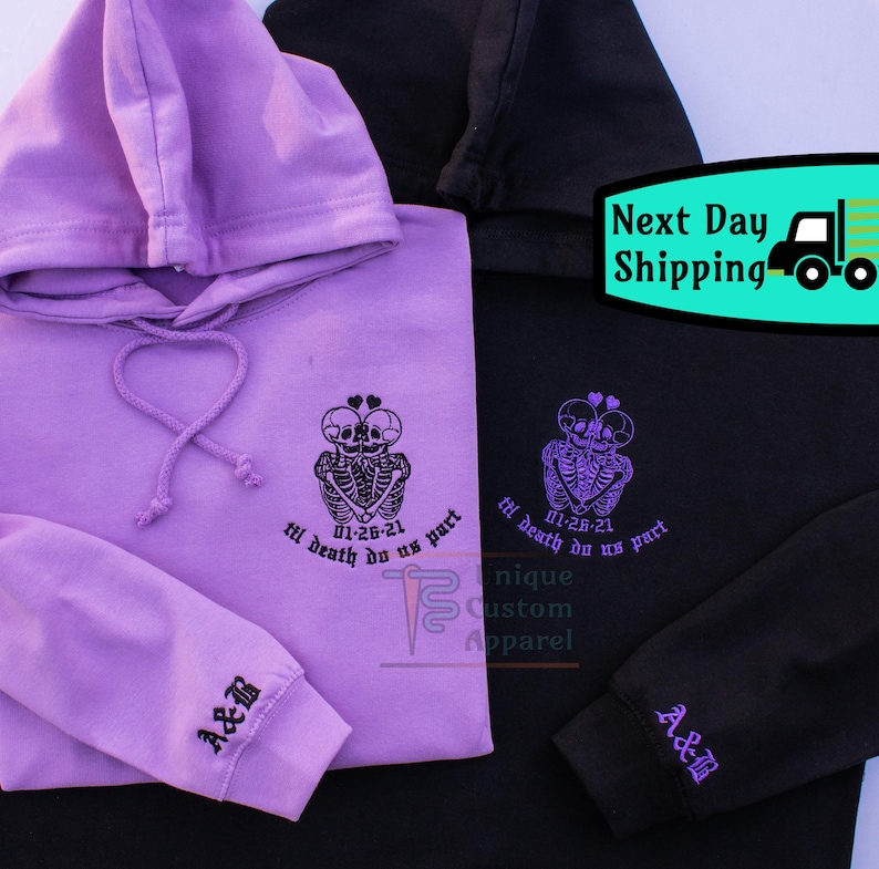 Two hoodies, one black and one lavender, are shown folded with the front of the black hoodie and the back of the lavender hoodie visible. Both have an embroidered design of two skeletons embracing with a heart above their heads, and the text "til
