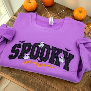 May include: A lavender long-sleeve sweatshirt featuring black embroidered varsity-style lettering that reads SPOOKY with white ghost and black bat accents. Below, the word season is embroidered in yellow cursive script.