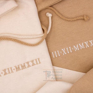 Op de afbeelding: Twee stoffen trekkoordtassen in beige en wit. De tassen zijn geborduurd met de Romeinse datum "III-XII-MMXXII".