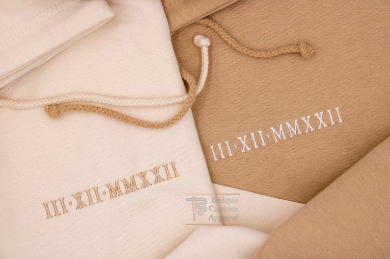 Op de afbeelding: Twee stoffen trekkoordtassen in beige en wit. De tassen zijn geborduurd met de Romeinse datum "III-XII-MMXXII".