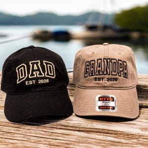 May include: A black baseball cap with the embroidered text DAD EST 2026 in gold and a tan baseball cap with the embroidered text GRANDPA EST 2026 in black. Both caps feature a curved brim and a structured crown with a small sticker on the tan cap.