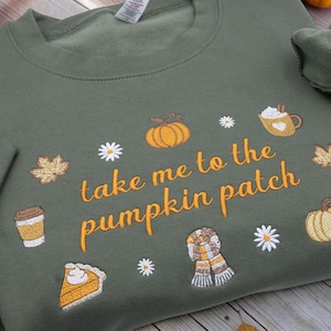 May include: An olive green sweatshirt featuring embroidered autumn-themed icons including pumpkins, maple leaves, daisies, a coffee cup, a slice of pumpkin pie, and a scarf, with the orange text "take me to the pumpkin patch" centred.