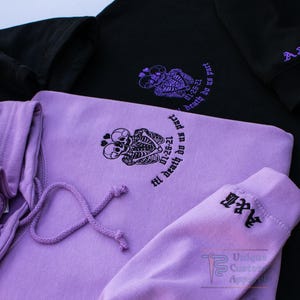 Two hoodies, one black and one lavender, are shown from an overhead angle, displaying the embroidered skeleton design and text on the front of both, and the embroidered initials on the sleeves.
