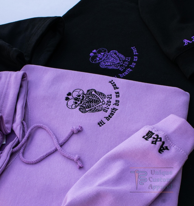 Two hoodies, one black and one lavender, are shown from an overhead angle, displaying the embroidered skeleton design and text on the front of both, and the embroidered initials on the sleeves.