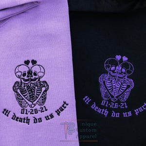 A close-up of two hoodies, one lavender and one black, showing the embroidered design of two skeletons embracing with a heart above their heads, and the text "til death do us part" and a date.