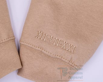 May include: A beige sweatshirt sleeve featuring embroidered Roman numerals XII VII XXI in a matching thread color. The fabric has a smooth, knit texture with a visible cuff seam. A small logo with the text Unique Custom Apparel is visible.