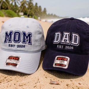 May include: A light blue baseball cap with the embroidered text MOM EST 2026 and a navy blue baseball cap with the embroidered text DAD EST 2026. Both caps feature a sticker on the brim with the brand name OTTO and the text DAD HAT.