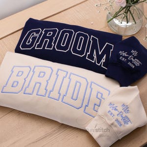 May include: Navy and cream sweatshirts and face coverings with embroidered text. The navy sweatshirt says "GROOM" in white block letters. The cream sweatshirt says "BRIDE" in blue block letters. Two face coverings are also visible, one navy and one cream, with custom text.