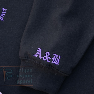 A close-up of the sleeve of a black hoodie, showing embroidered initials "A&B" in a gothic font.