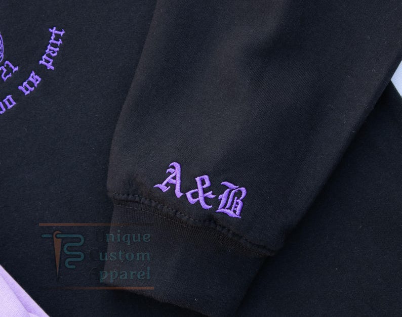 A close-up of the sleeve of a black hoodie, showing embroidered initials "A&B" in a gothic font.