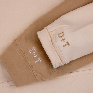 Op de afbeelding: Close-up van twee sweatshirtmouwen in bruin en cr&egrave;me met de tekst "D+T" geborduurd op elke mouw.
