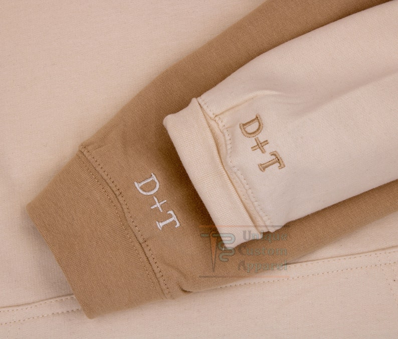 Op de afbeelding: Close-up van twee sweatshirtmouwen in bruin en cr&egrave;me met de tekst "D+T" geborduurd op elke mouw.