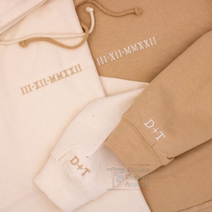 Op de afbeelding: Twee hoodies, een beige en een cr&egrave;mekleurige, met de tekst "III-XII-MMXXII" geborduurd op de voorkant en "D+T" geborduurd op de mouw.