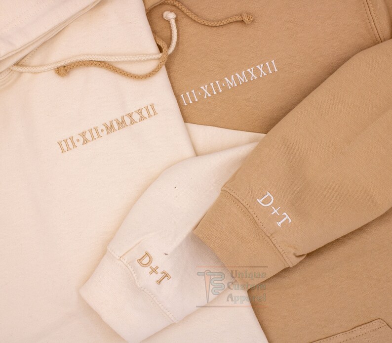 Op de afbeelding: Twee hoodies, een beige en een cr&egrave;mekleurige, met de tekst "III-XII-MMXXII" geborduurd op de voorkant en "D+T" geborduurd op de mouw.