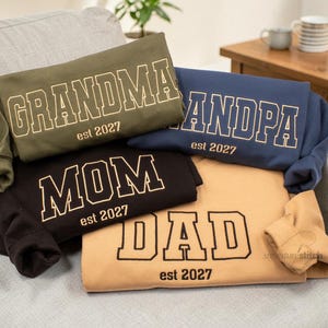 May include: Four folded sweatshirts in olive green, navy blue, black, and tan. Each features collegiate-style lettering reading GRANDMA, GRANDPA, MOM, or DAD, with the text est 2027 printed below. The garments have long sleeves and a crew neckline.