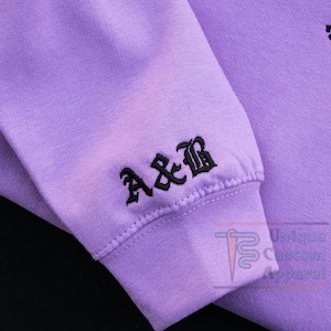 A close-up of the sleeve of a lavender hoodie, showing embroidered initials "A&B" in a gothic font.