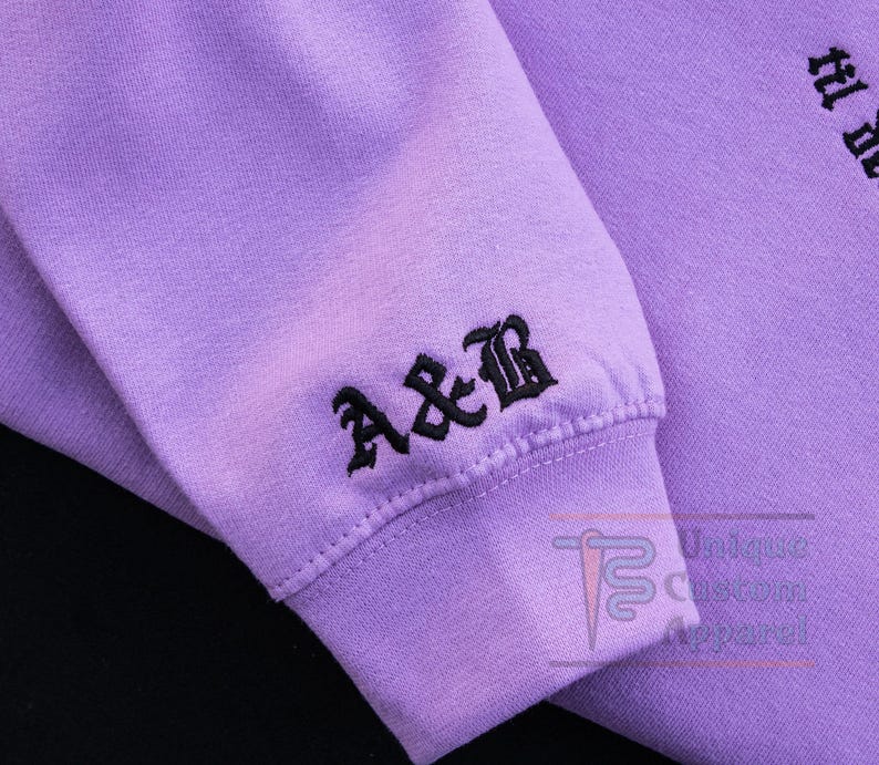 A close-up of the sleeve of a lavender hoodie, showing embroidered initials "A&B" in a gothic font.
