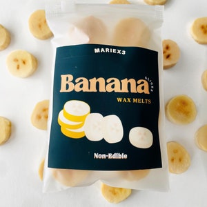 May include: A clear plastic bag with a label that says "Banana Slices Wax Melts" and "Non-Edible". The bag contains several wax melts shaped like banana slices. The wax melts are a light yellow color.