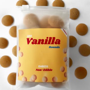 Op de afbeelding: Een doorzichtige plastic zak gevuld met kleine, ronde, bruine, vanillegeurende wax melts. De zak heeft een geel label met de tekst "MINI vanilla Rounds EMBEDS Non-Edible".
