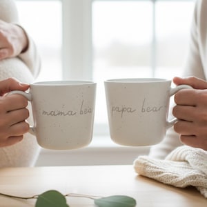 May include: Two off-white ceramic mugs with a speckled finish, each featuring a handle. One mug has the text "mama bear" in a thin, dark script font, and the other mug has the text "papa bear" in the same matching dark script font style.
