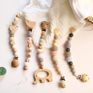 May include: A collection of wooden baby pacifier clips with various animal shapes and bead designs. The clips feature a bear, bird, elephant, and heart. The beads are in neutral colors, including beige, gray, and pastel shades.