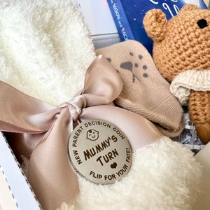 May include: A white gift box with a pink ribbon and a wooden coin that says "New Parent Decision Coin, Mummy's Turn, Flip for Your Fate!" The box contains a white fuzzy sock, a beige sock with a star pattern, and a crocheted brown bear with a white crocheted scarf.