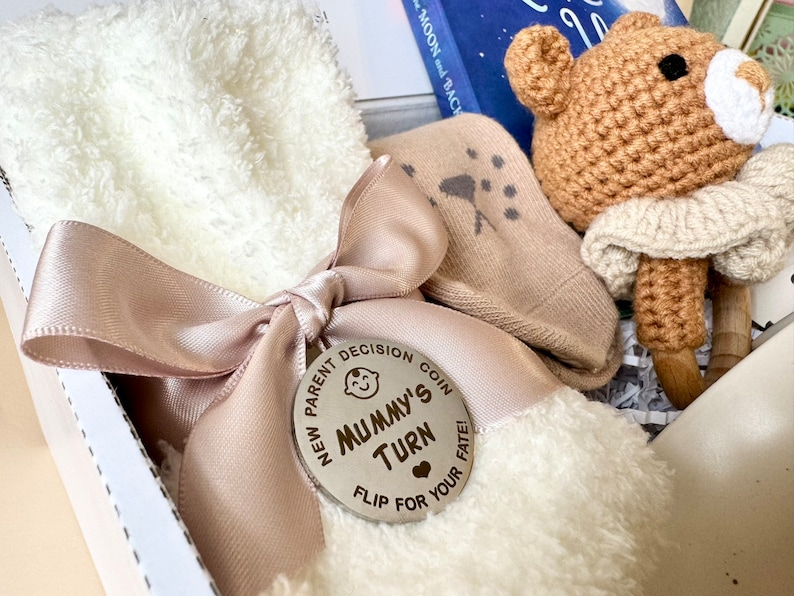 May include: A white gift box with a pink ribbon and a wooden coin that says "New Parent Decision Coin, Mummy's Turn, Flip for Your Fate!" The box contains a white fuzzy sock, a beige sock with a star pattern, and a crocheted brown bear with a white crocheted scarf.