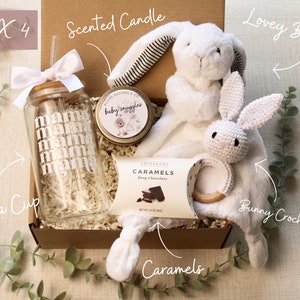 May include: A gift box filled with baby items, including a white crocheted bunny rattle, a white lovey bunny, a glass cup with the word "mama" repeated on it, a small jar of scented candle, and a box of caramels. The box is labeled "BOX 4".
