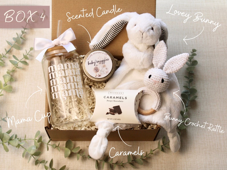 May include: A gift box filled with baby items, including a white crocheted bunny rattle, a white lovey bunny, a glass cup with the word "mama" repeated on it, a small jar of scented candle, and a box of caramels. The box is labeled "BOX 4".