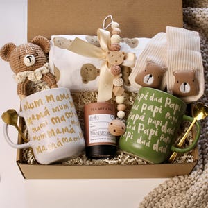 Könnte beinhalten: Eine Baby-Geschenkbox mit einem gehäkelten Teddybären, zwei Tassen mit goldenen Griffen und Text, einer Babydecke, einem Holzbeißring und einer Teedose. Auf den Tassen steht "Mum Mama" und "Papi Dad".