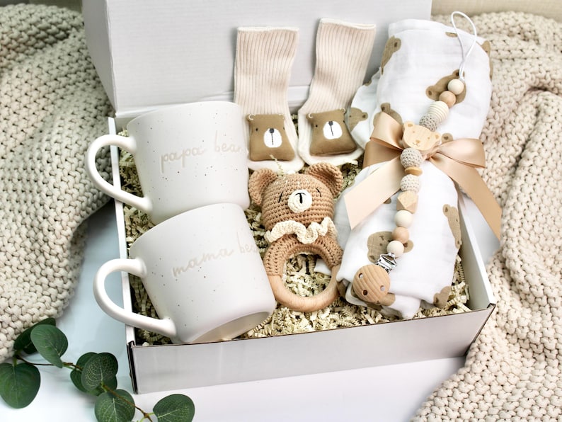 Peut inclure: Une bo&icirc;te cadeau blanche remplie d'un ensemble cadeau de f&ecirc;te pr&eacute;natale. L'ensemble comprend deux tasses blanches avec le texte "papa bear" et "mama bear", un hochet en crochet en forme d'ours brun, une couverture en mousseline blanche &agrave; imprim&eacute; ours brun et un anneau de dentition en perles de bois avec un clip.