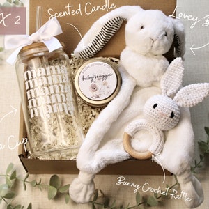 May include: A gift box filled with baby items, including a white crocheted bunny rattle, a white lovey bunny, a glass cup with the word "mama" repeated on it, and a small candle with the words "baby snuggles".
