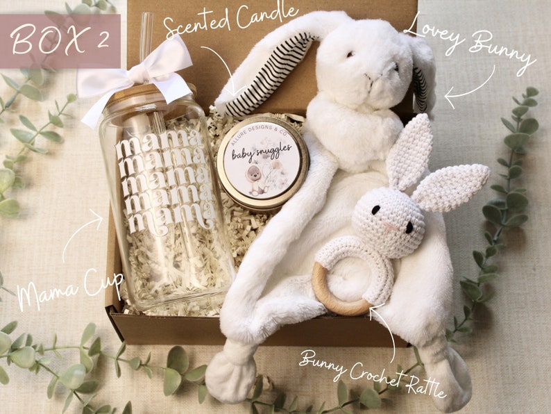 May include: A gift box filled with baby items, including a white crocheted bunny rattle, a white lovey bunny, a glass cup with the word "mama" repeated on it, and a small candle with the words "baby snuggles".