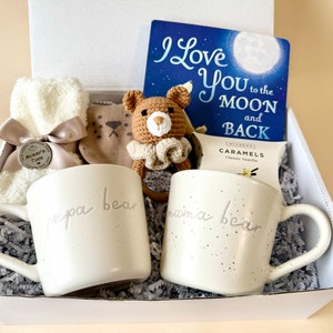 May include: A white gift box filled with a variety of items, including two white mugs with the words "papa bear" and "mama bear" on them, a brown crocheted bear toy, a book titled "I Love You to the Moon and Back", a pair of white fuzzy socks, and a small bag of caramels.
