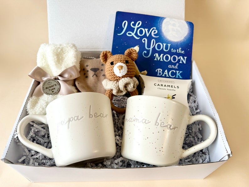 May include: A white gift box filled with a variety of items, including two white mugs with the words "papa bear" and "mama bear" on them, a brown crocheted bear toy, a book titled "I Love You to the Moon and Back", a pair of white fuzzy socks, and a small bag of caramels.
