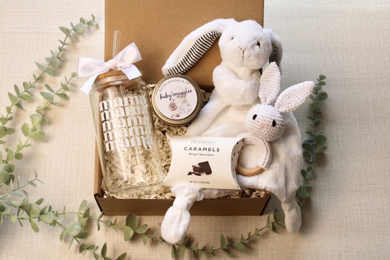 May include: A gift box filled with items for a new mom. The box contains a glass water bottle with a straw, a small tin of candle, a box of chocolates, and a white plush bunny with a crocheted bunny attached.