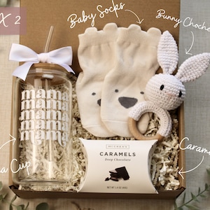 May include: A gift box filled with baby items, including a white crocheted bunny rattle, a pair of baby socks, a glass cup with the word "mama" printed on it, and a package of chocolate caramels.